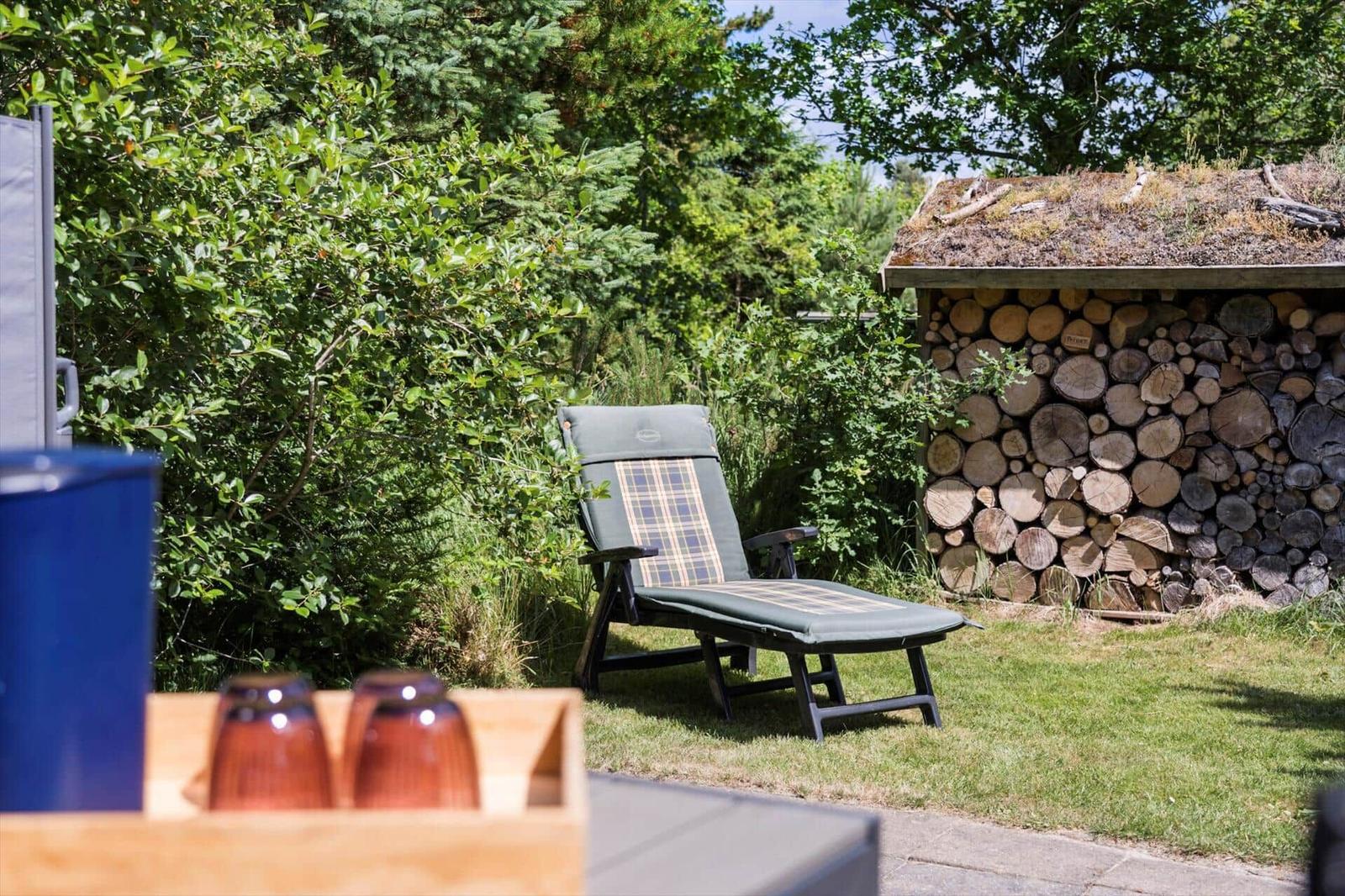 Liegestuhl im Garten neben Holzstapel und grüner Vegetation.