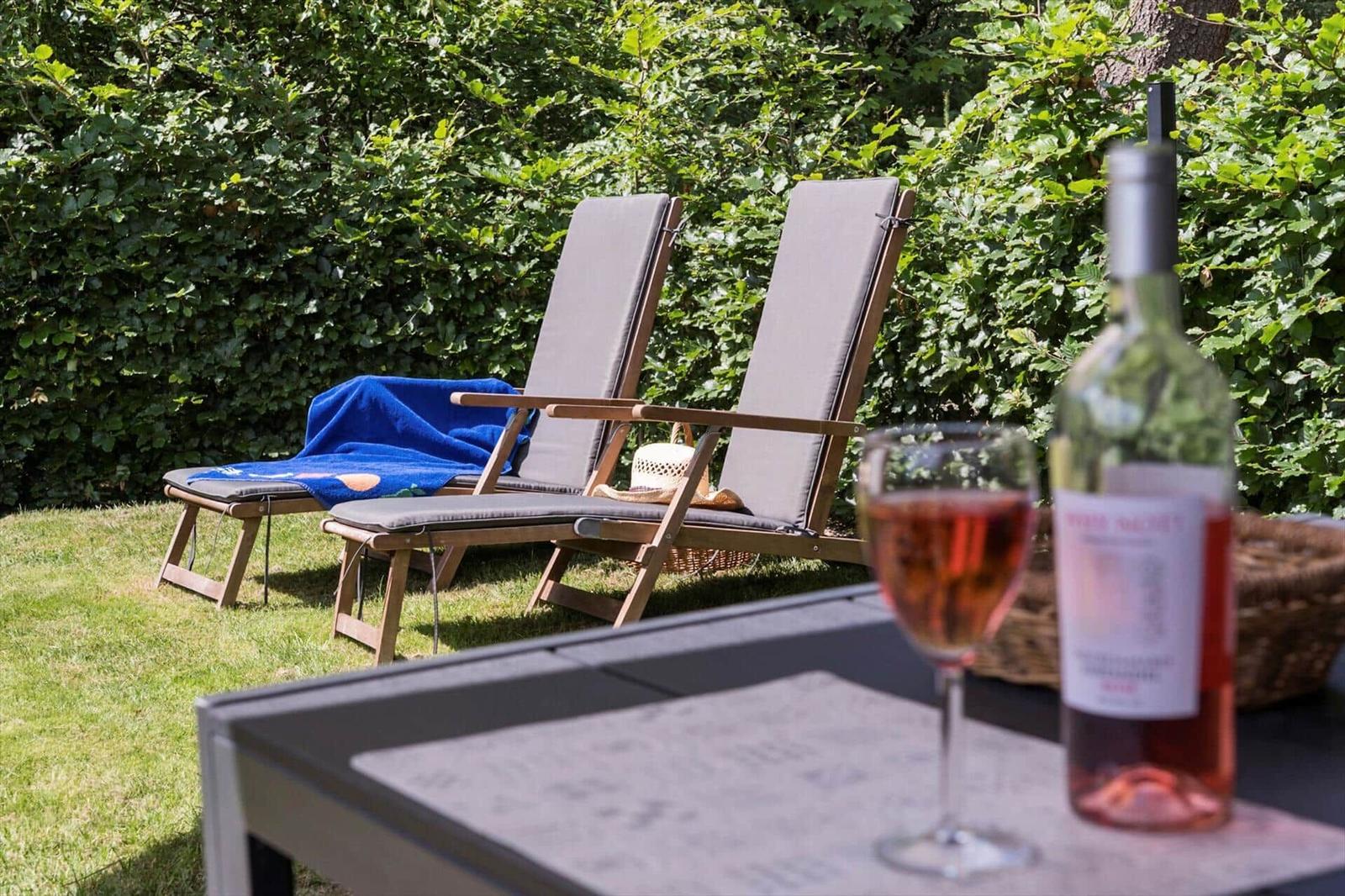 Zwei Liegestühle mit blauer Decke stehen im Garten. Auf Tisch steht ein Glas und eine Flasche Rosé.