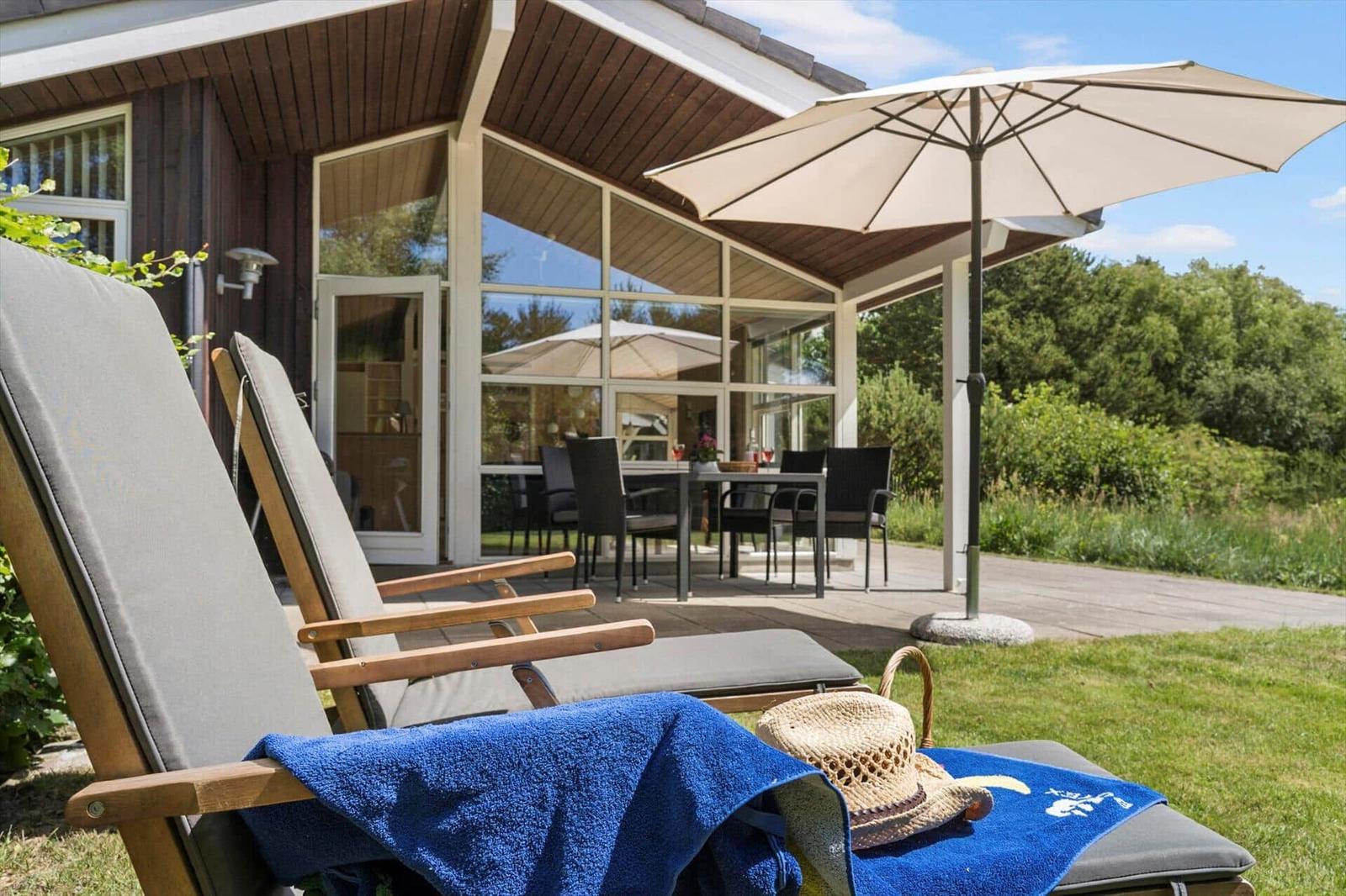 Terrasse mit Liegestühlen, Sonnenschirm und Tisch. Hintergrund: Haus mit Glasfront und Grün.
