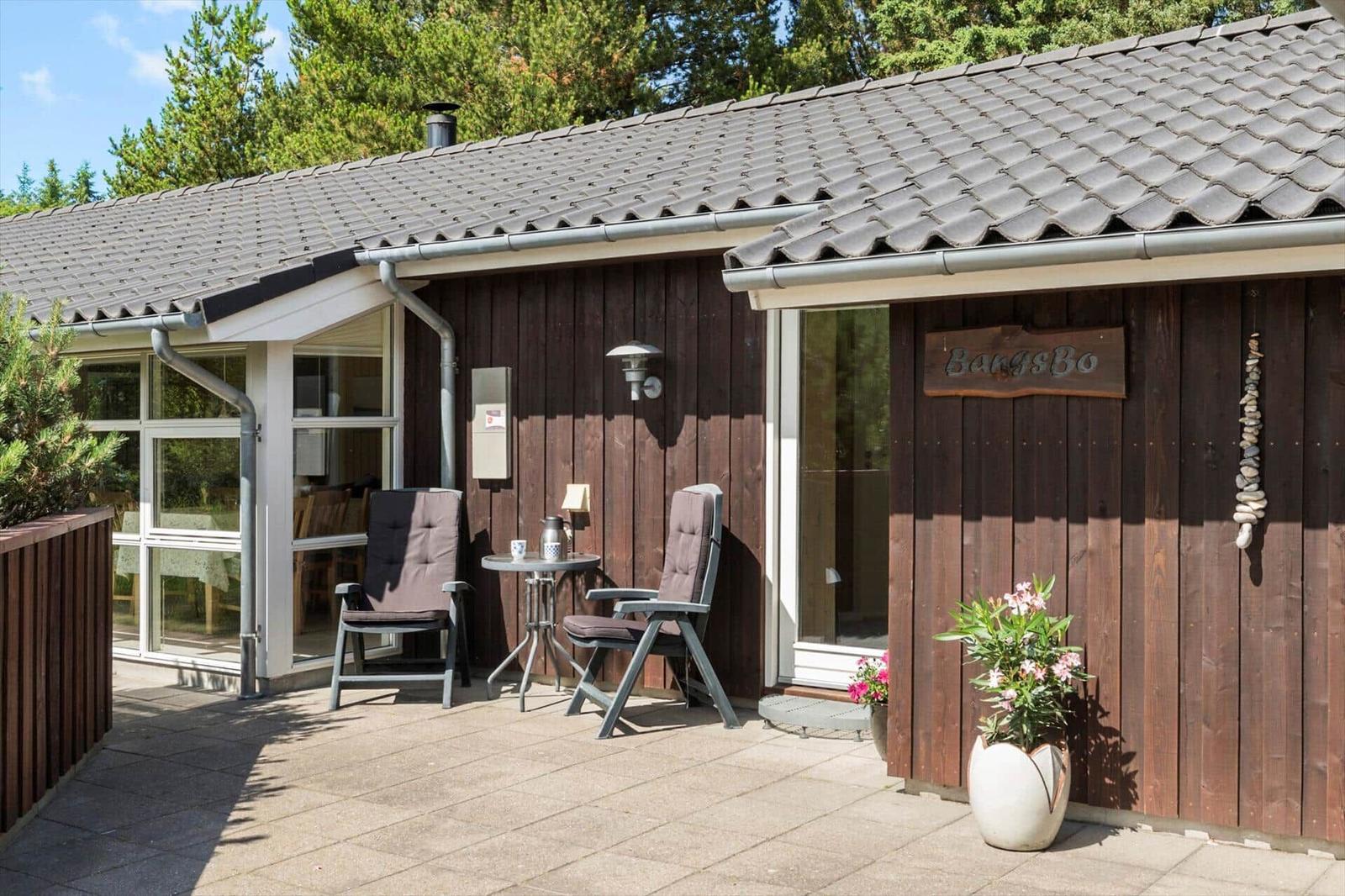 Haus mit Holzfassade, Terrasse mit zwei Stühlen und Tisch, Pflanze vor der Tür.