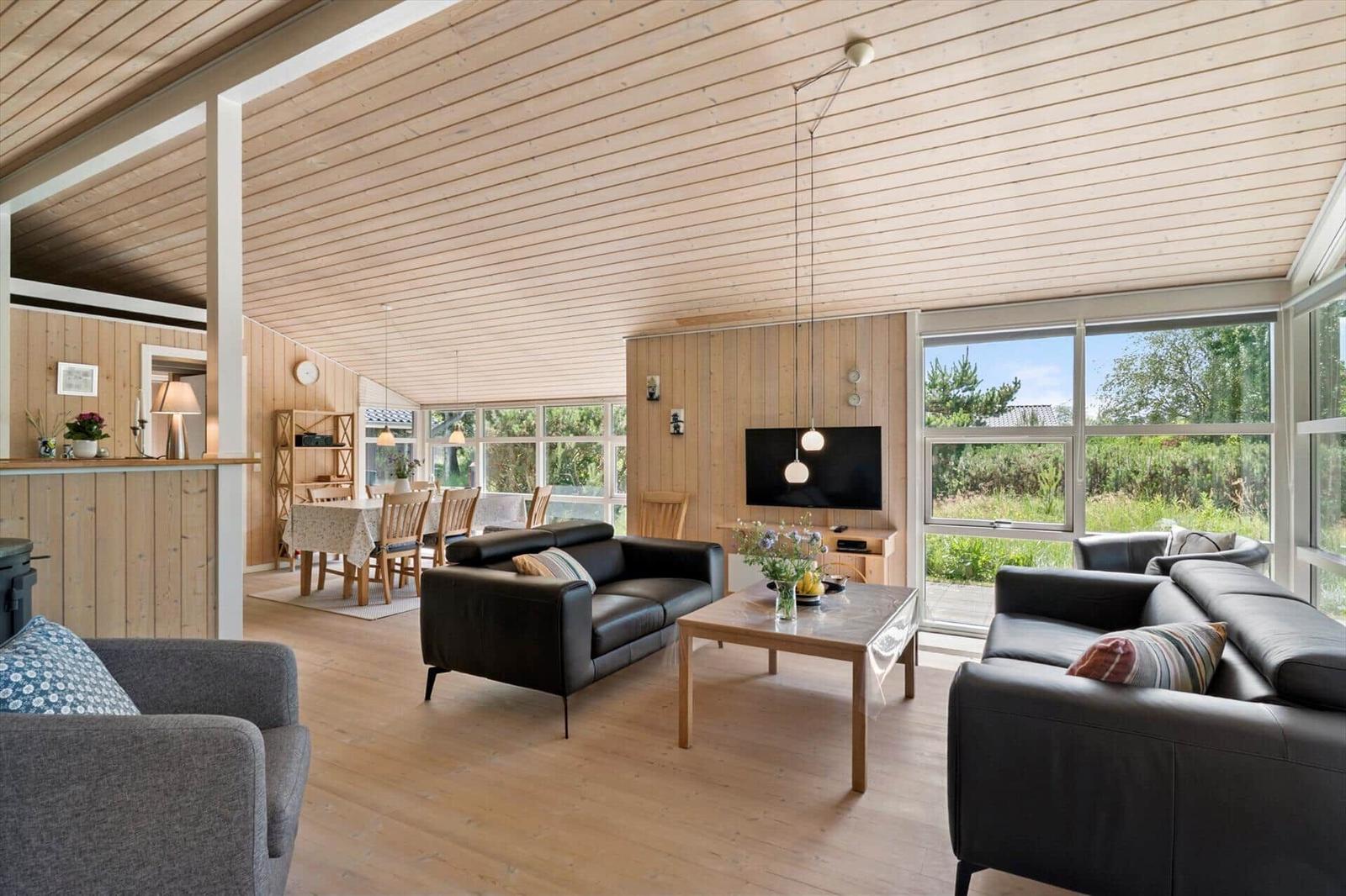 Wohn- und Essbereich mit Holzdecke, schwarzen Sofas und großen Fenstern.