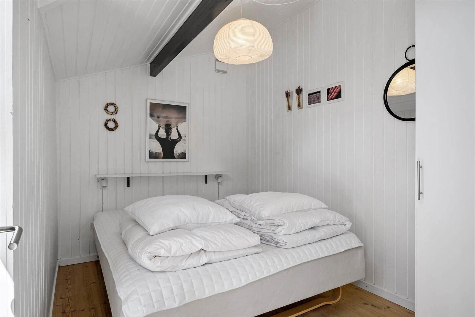 Schlafzimmer mit weißem Bett, Holzboden und weißen Wänden. Deckenbalken und Pendelleuchte.