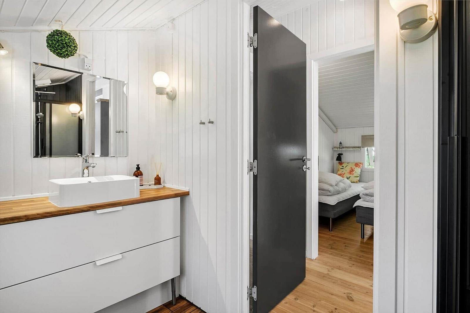 Badezimmer mit Waschtisch und Spiegel. Tür führt in Schlafzimmer mit Bett.