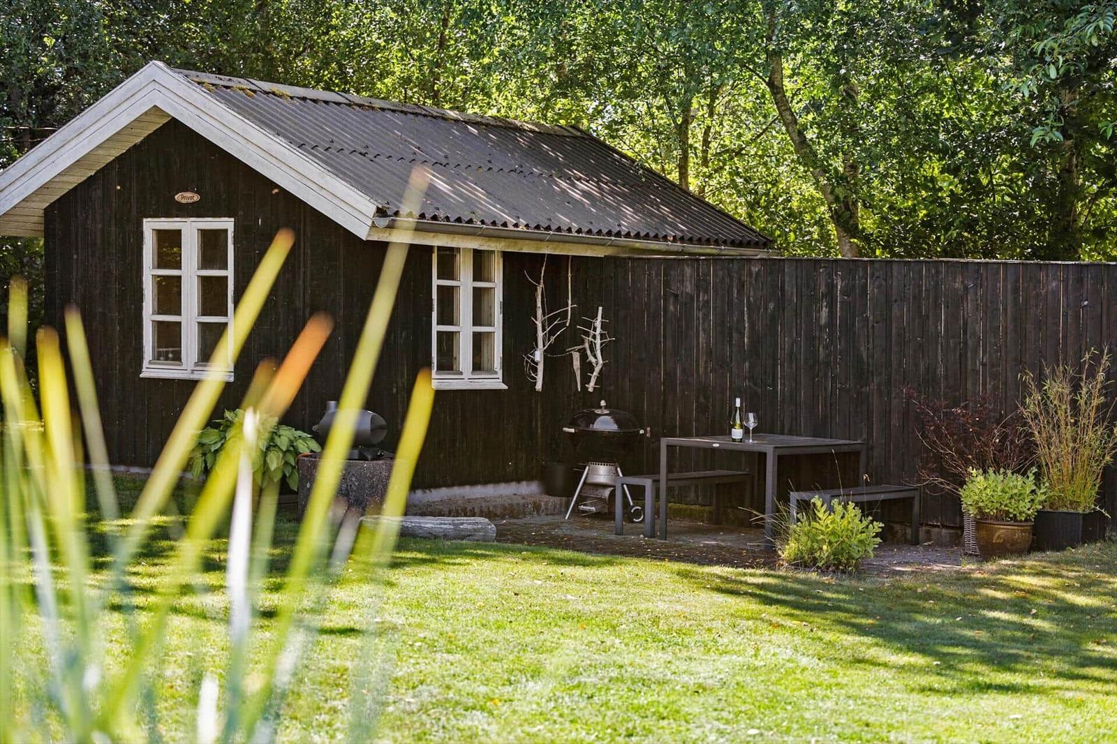 Hütte mit Garten, Grill und Tisch. Umgeben von Bäumen und Gras.