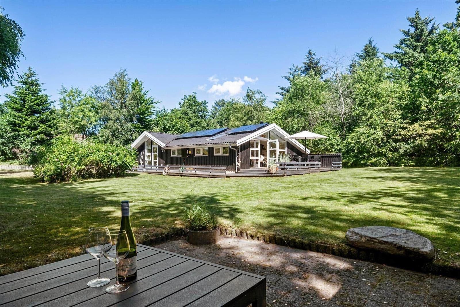 Holzhütte mit Terrasse, Garten und Sonnenkollektoren. Tisch mit Wein im Vordergrund.