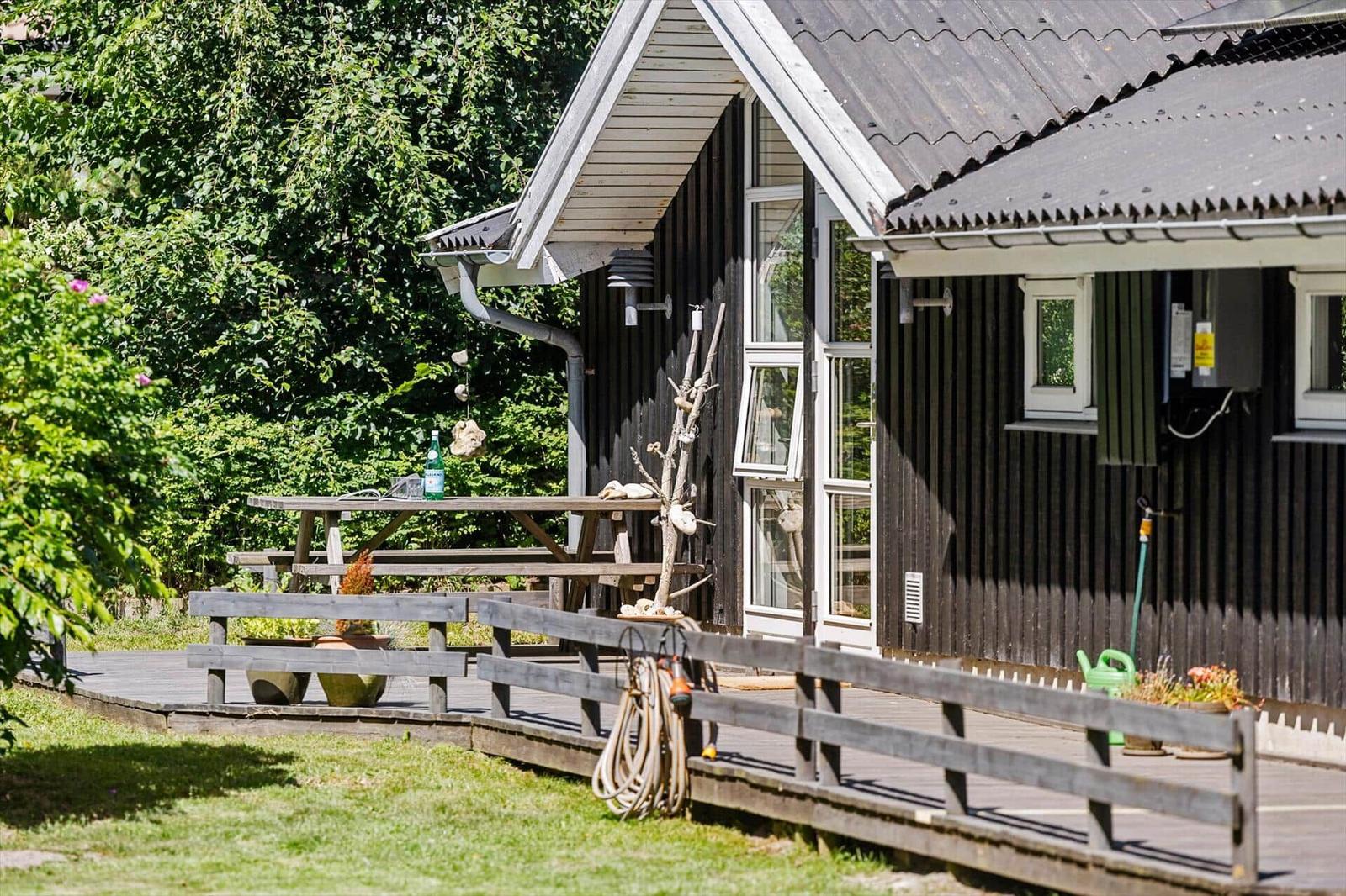 Holzterrasse mit Tisch, Gartenmöbeln und Pflanzen vor einem Haus mit dunkler Fassade.