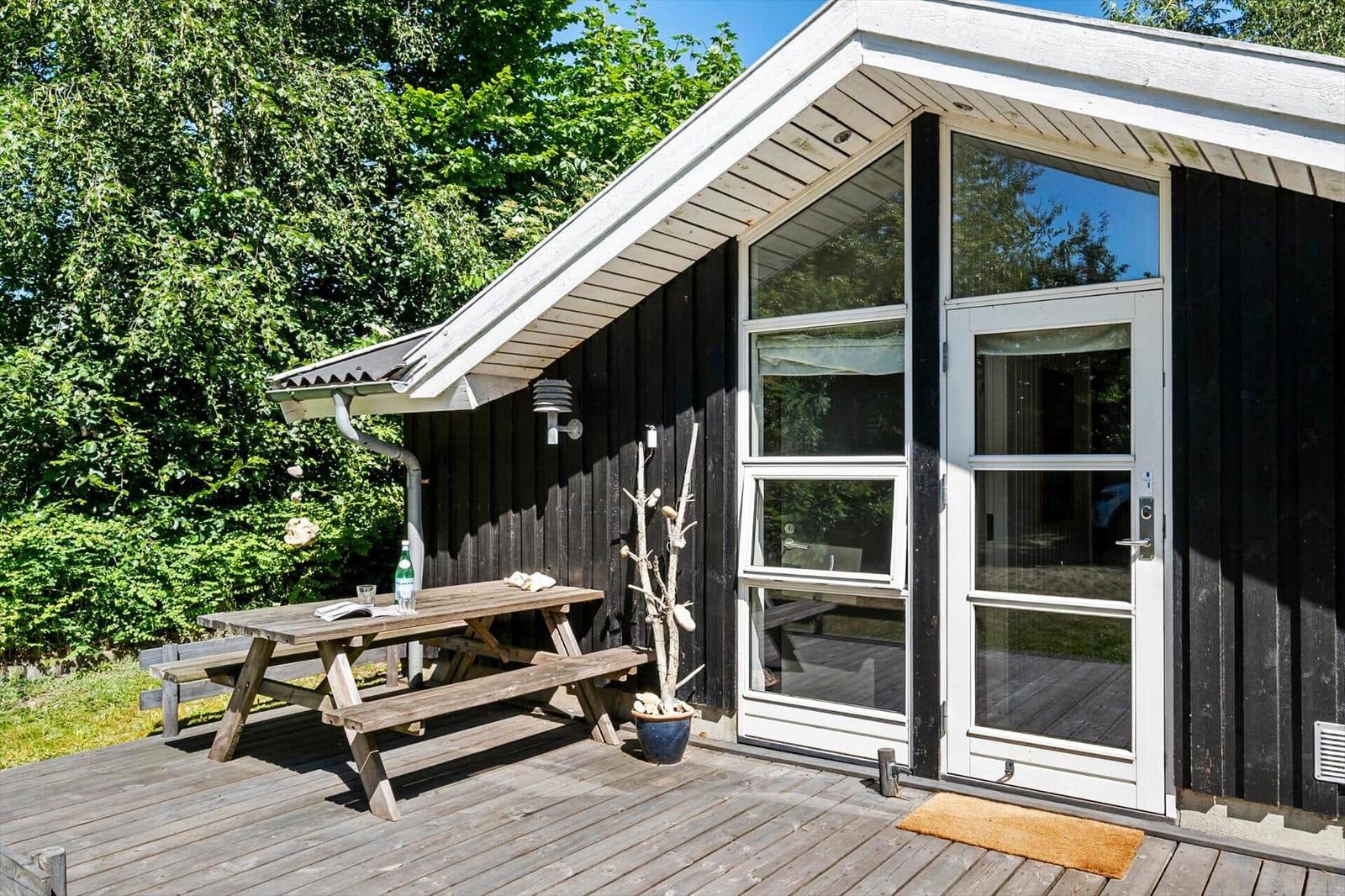 Hütte mit Holzterrasse, Picknicktisch und Glastür. Umgeben von Bäumen.