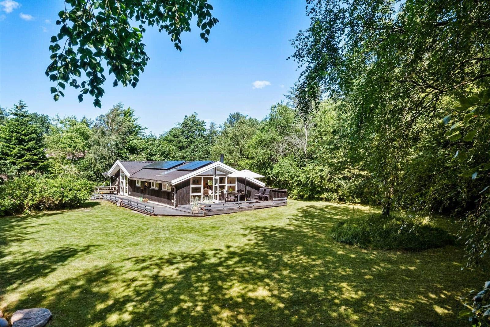 Ein Holzhaus mit Terrasse und Solaranlage auf grünem Grundstück umgeben von Bäumen.