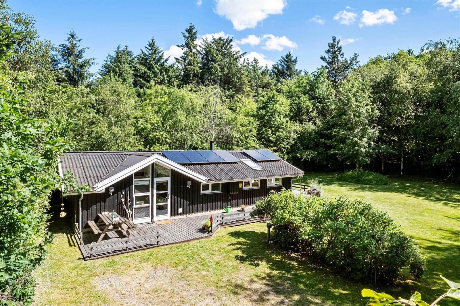 Holzhütte mit Solaranlage, Terrasse und Garten. Umgeben von Wald und Grün.