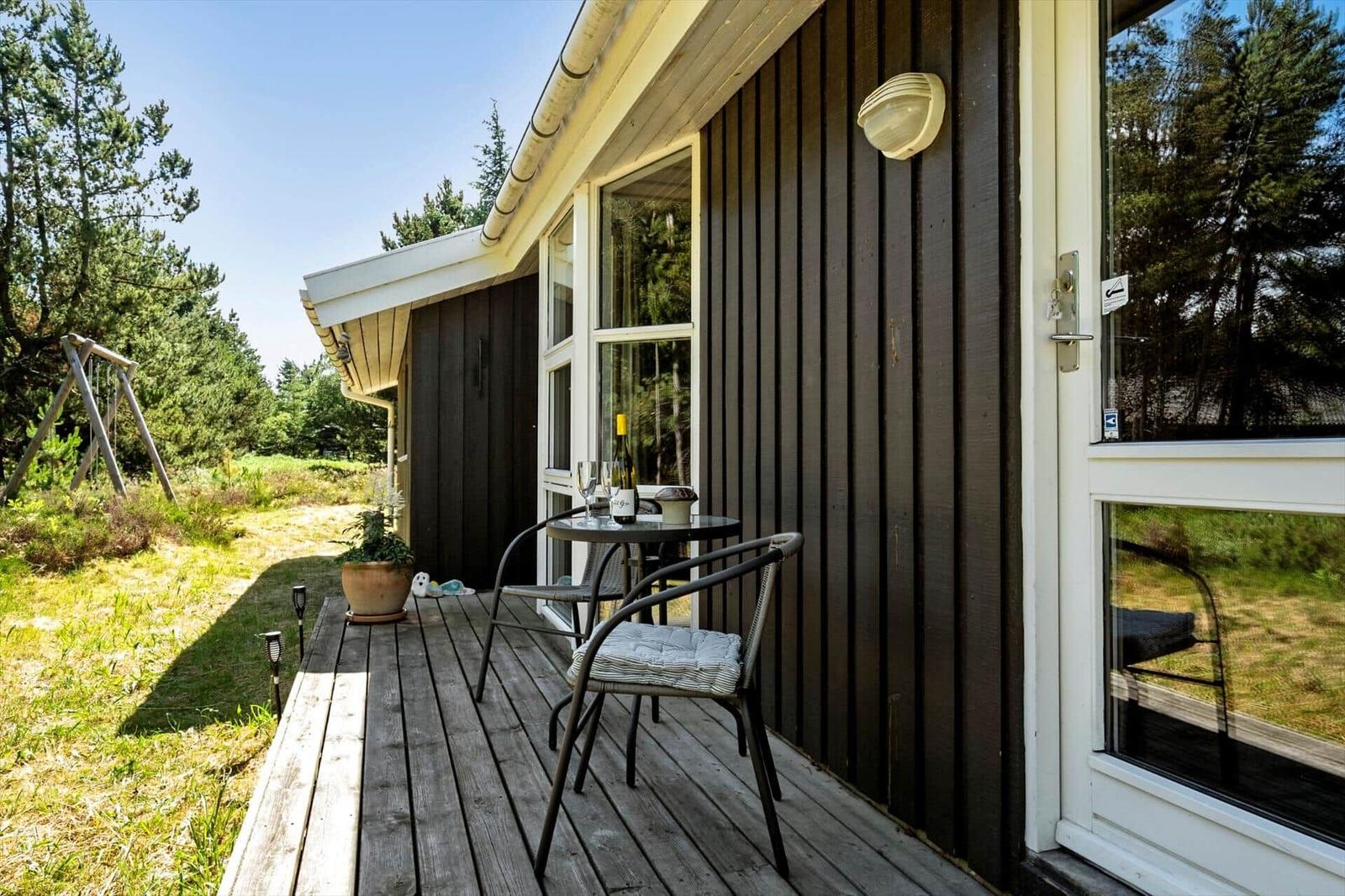 Terrasse mit Tisch, zwei Stühlen und Weinflasche vor einem Holzhaus mit Fenster und Tür.