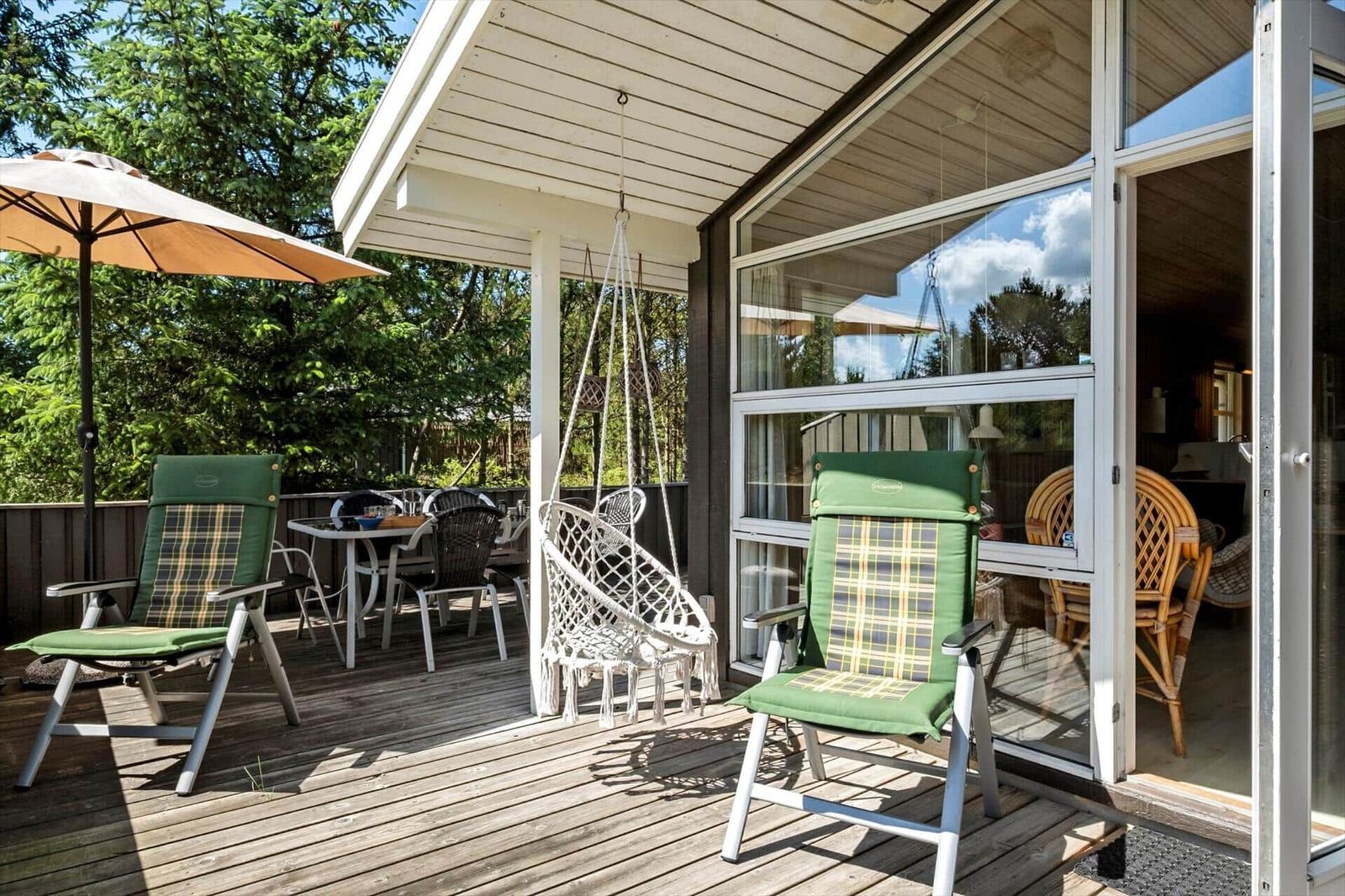Terrasse mit grünen Stühlen, Sonnenschirm und Hängesessel. Durchgehende Glasfront zum Innenraum.
