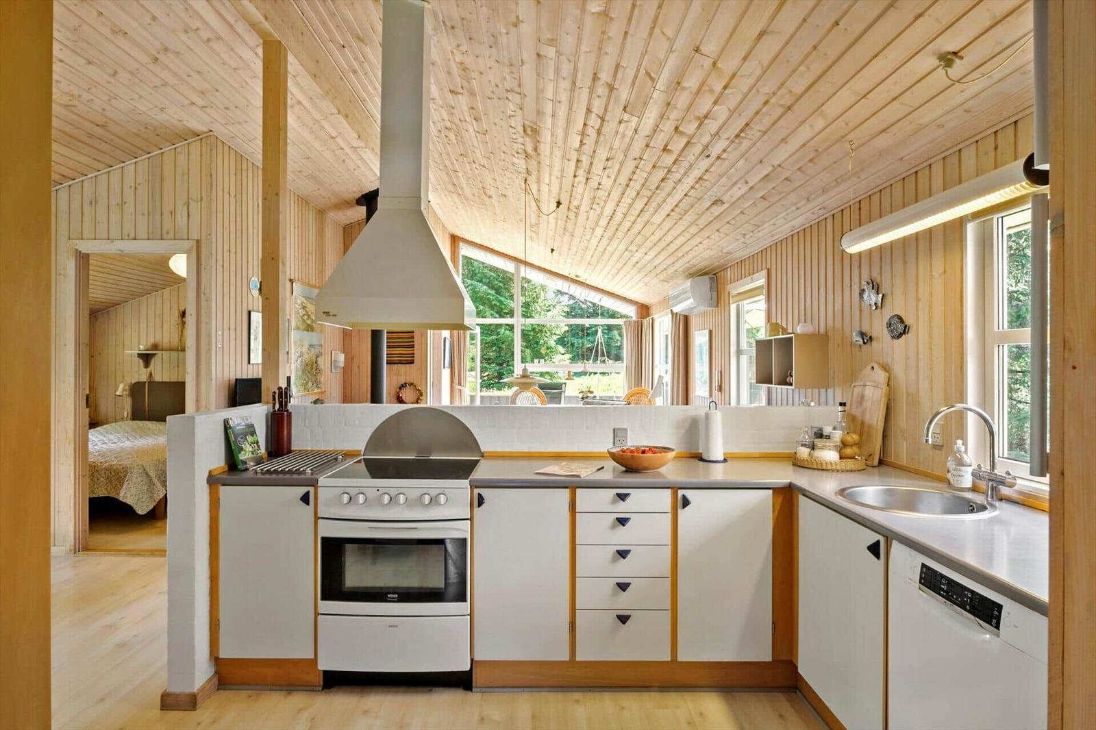 Küche mit weißen Schränken, Steinplatten und Holzverkleidung. Blick in Schlafzimmer.
