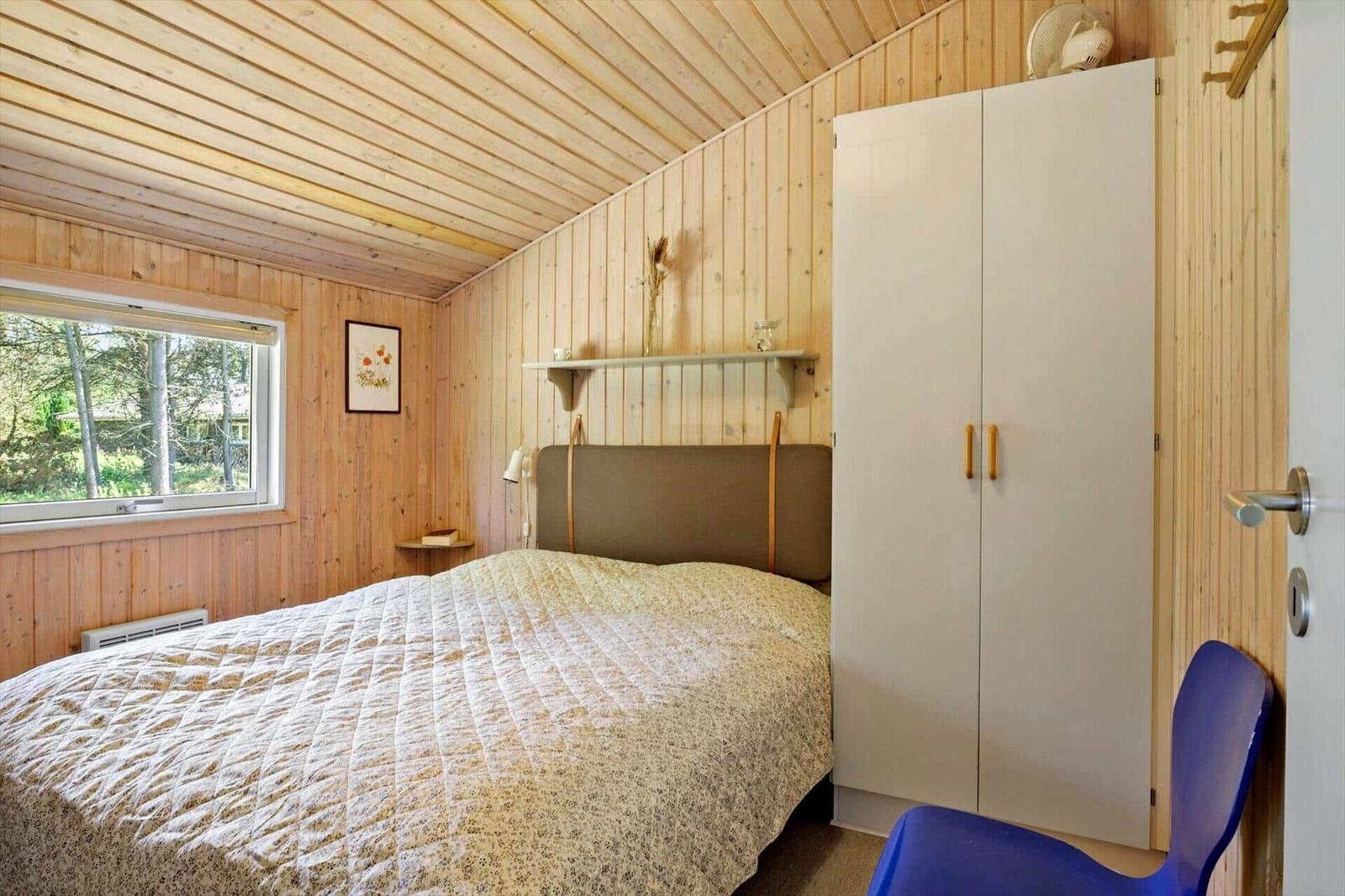 Schlafzimmer mit Holzwand, Bett, Kleiderschrank und Fenster mit Blick auf Wald.