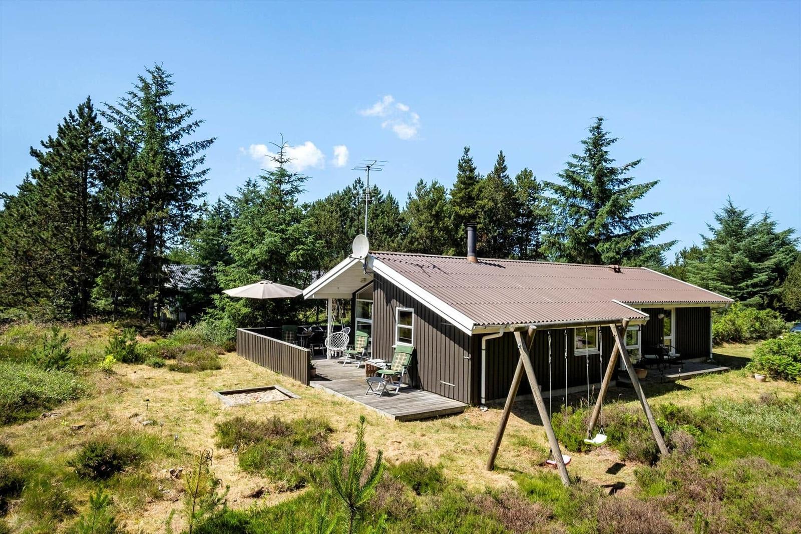 Holzhaus mit Terrasse, Schaukel und Garten. Umgeben von Nadelbäumen unter blauem Himmel.