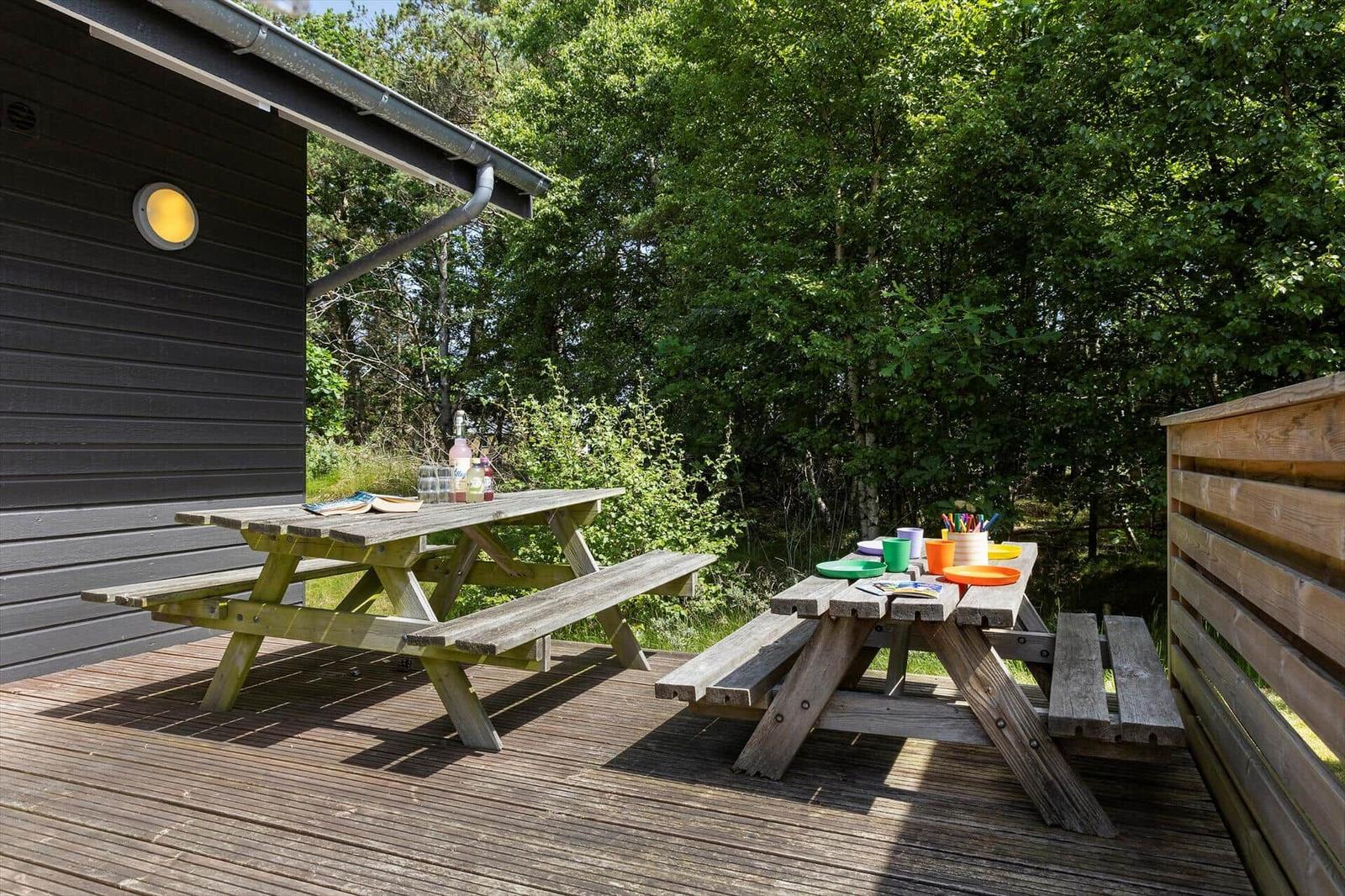 Holzterrasse mit zwei Picknicktischen, umgeben von Bäumen und einem dunklen Holzhaus.