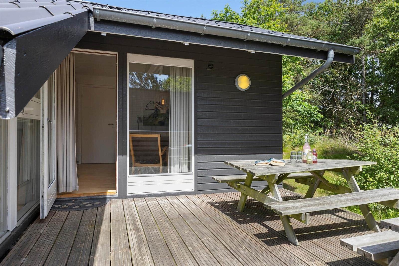 Terrasse mit Picknicktisch vor einem dunklen Holzhaus. Durch Fenster und Tür ist Innenraum sichtbar.