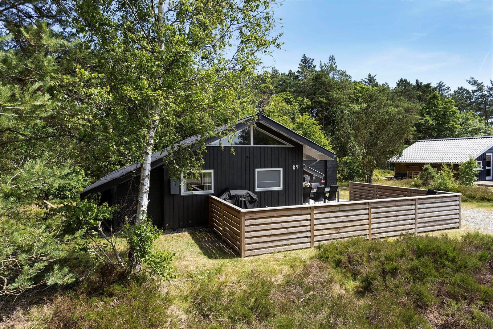 Schwarzes Ferienhaus mit Holzterrasse und Grill, umgeben von Bäumen und Grün.