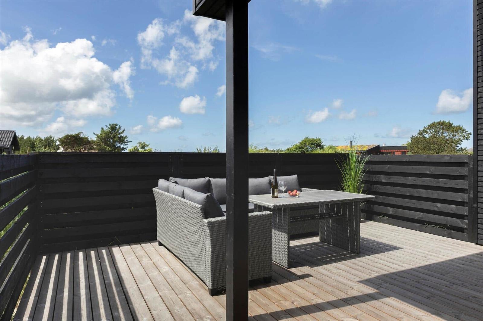 Terrasse mit Sitzgruppe, Tisch und Flasche. Holzdecke, schwarze Umzäunung, Himmel mit Wolken.