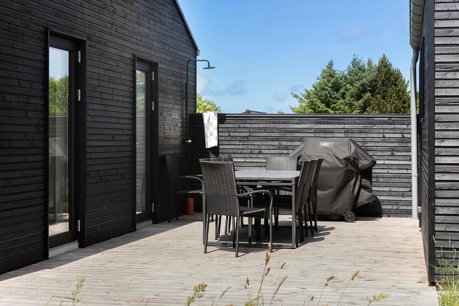 Terrasse mit Tisch, Stühlen und Grill, angrenzend an ein schwarz verkleidetes Haus.