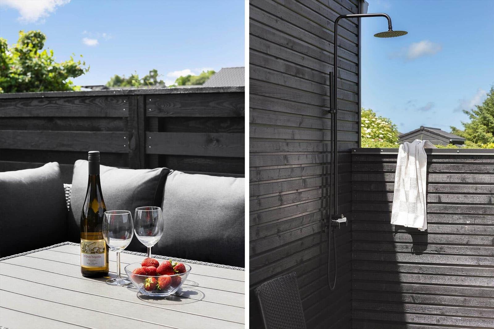 Terrasse mit Tisch, Stühlen, Wein und Erdbeeren. Dusche im Freien an Holzwand.