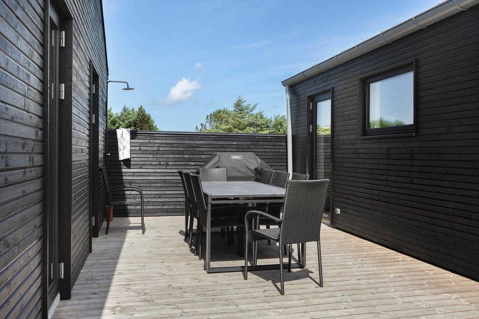 Terrasse mit Tisch, Stühlen und Grill. Holzwand und Dachrinnen sichtbar.