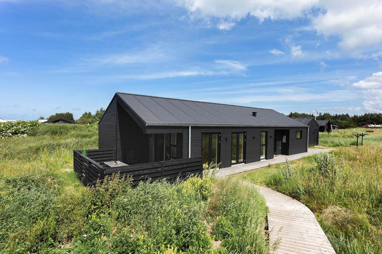 Moderne schwarze Hütte mit Holzterrasse und Weg durch Grasland unter blauem Himmel.