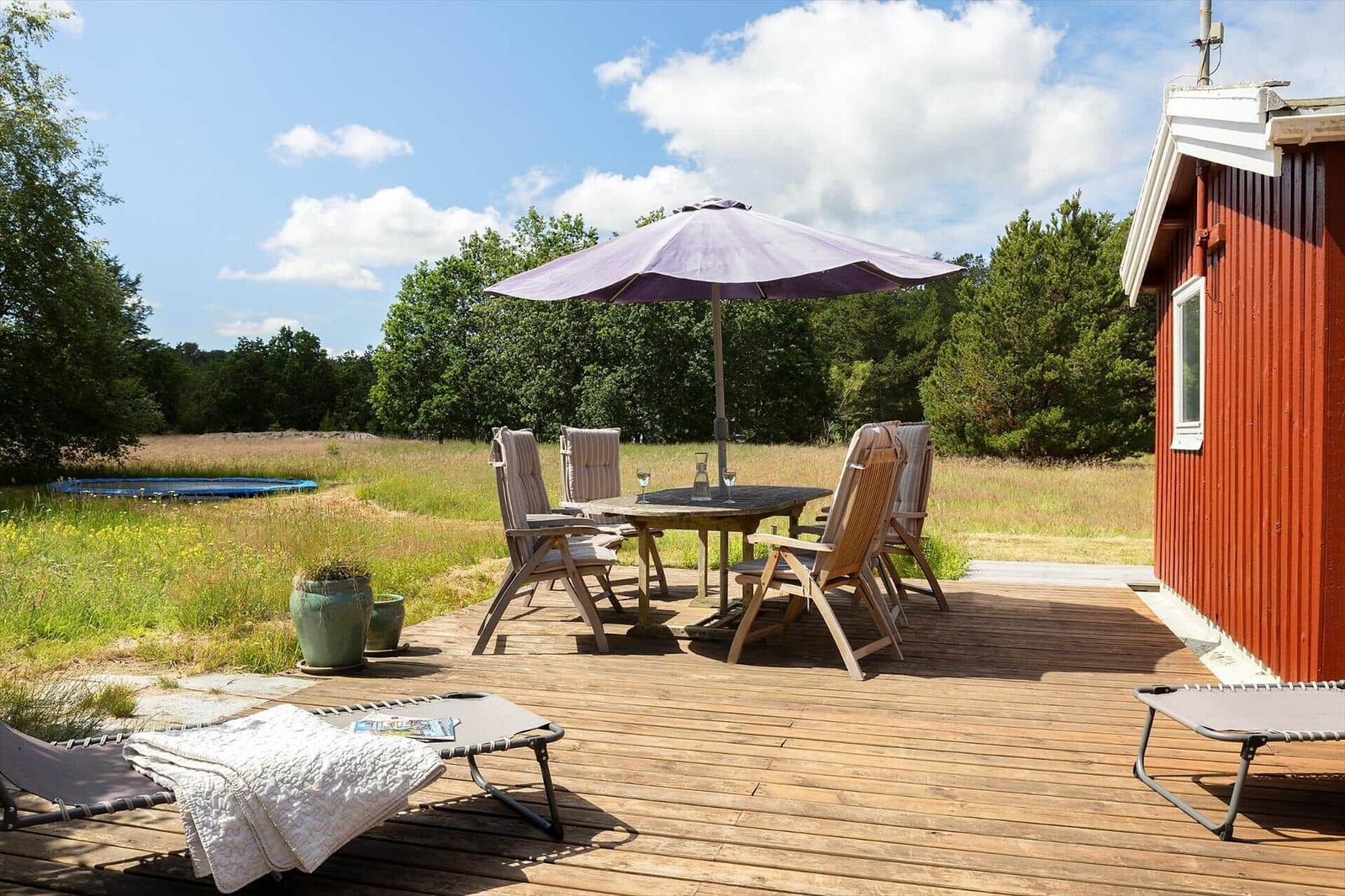 Holzterrasse mit Tisch, Stühlen und Sonnenschirm. Roter Holzschuppen und Grünfläche im Hintergrund.