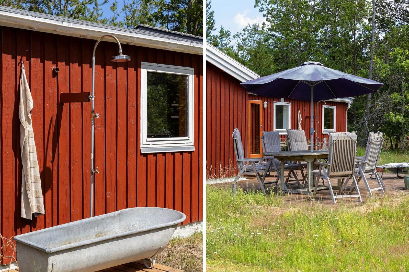Roter Holzhaus mit Außendusche und Badewanne. Terrasse mit Tisch, Stühlen und Sonnenschirm.