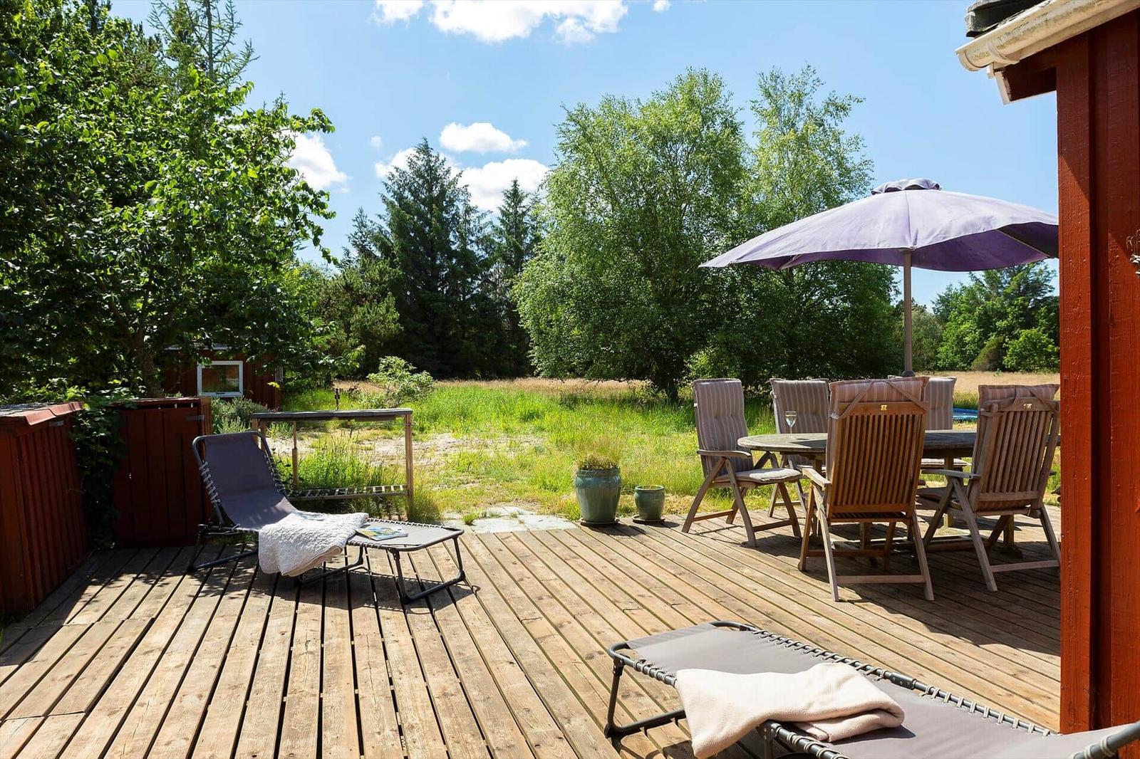 Holzterrasse mit Liegestühlen, Tisch und Sonnenschirm. Grünflächen und Bäume im Hintergrund.
