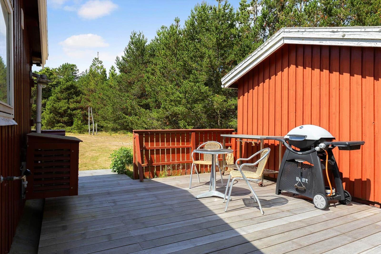 Terrasse mit Tisch, Stühlen und Grill neben rotem Holzhaus. Hintergrund: Wald.