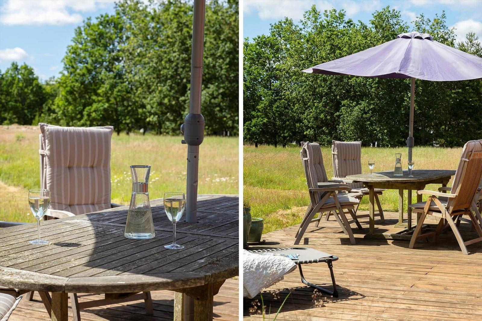 Terrasse mit Tisch, Stühlen und Sonnenschirm. Hintergrund: Grasland und Bäume.