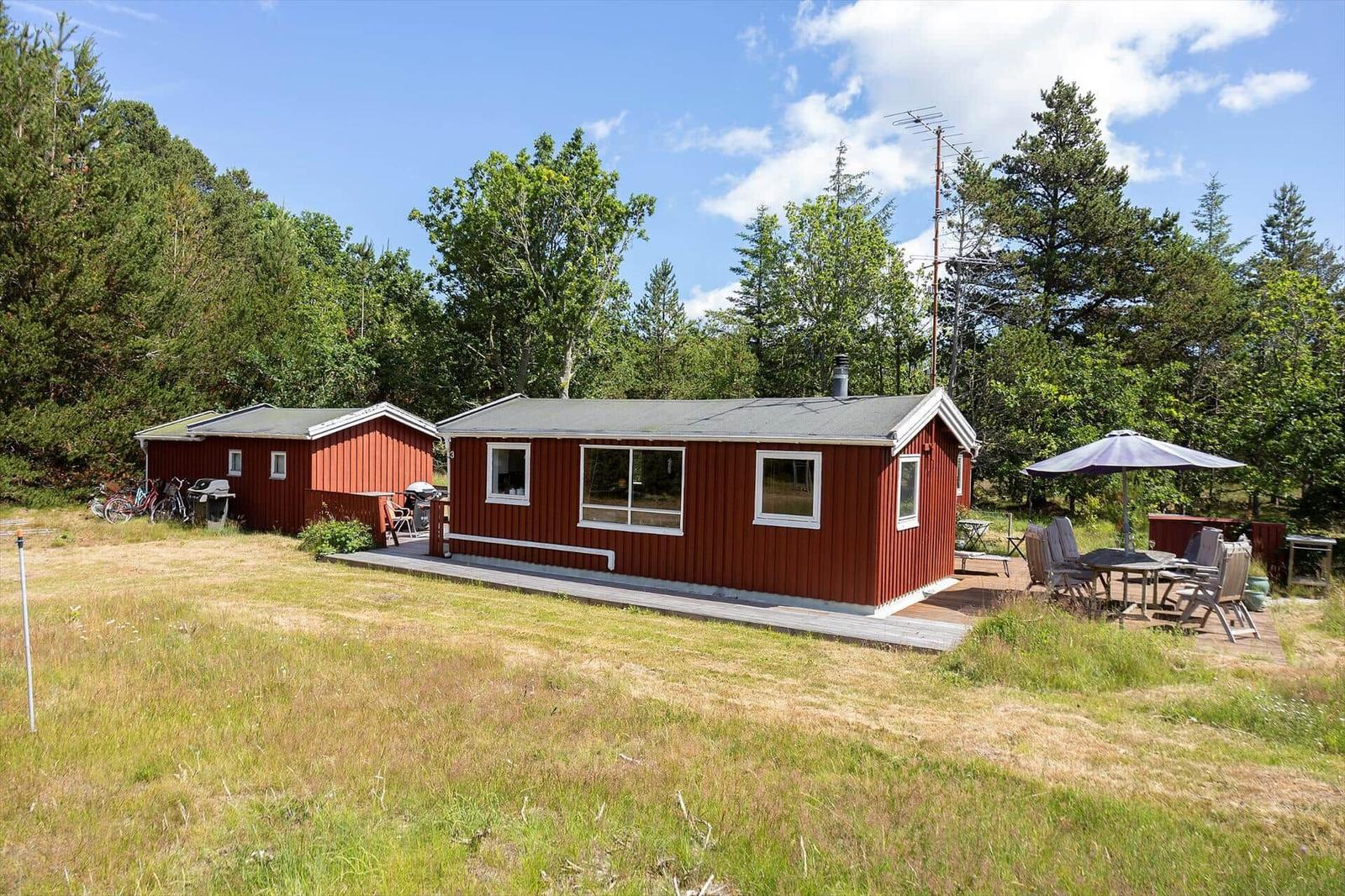 Rote Holzhütte mit Terrasse und Garten. Umgeben von Bäumen und einem klaren Himmel.