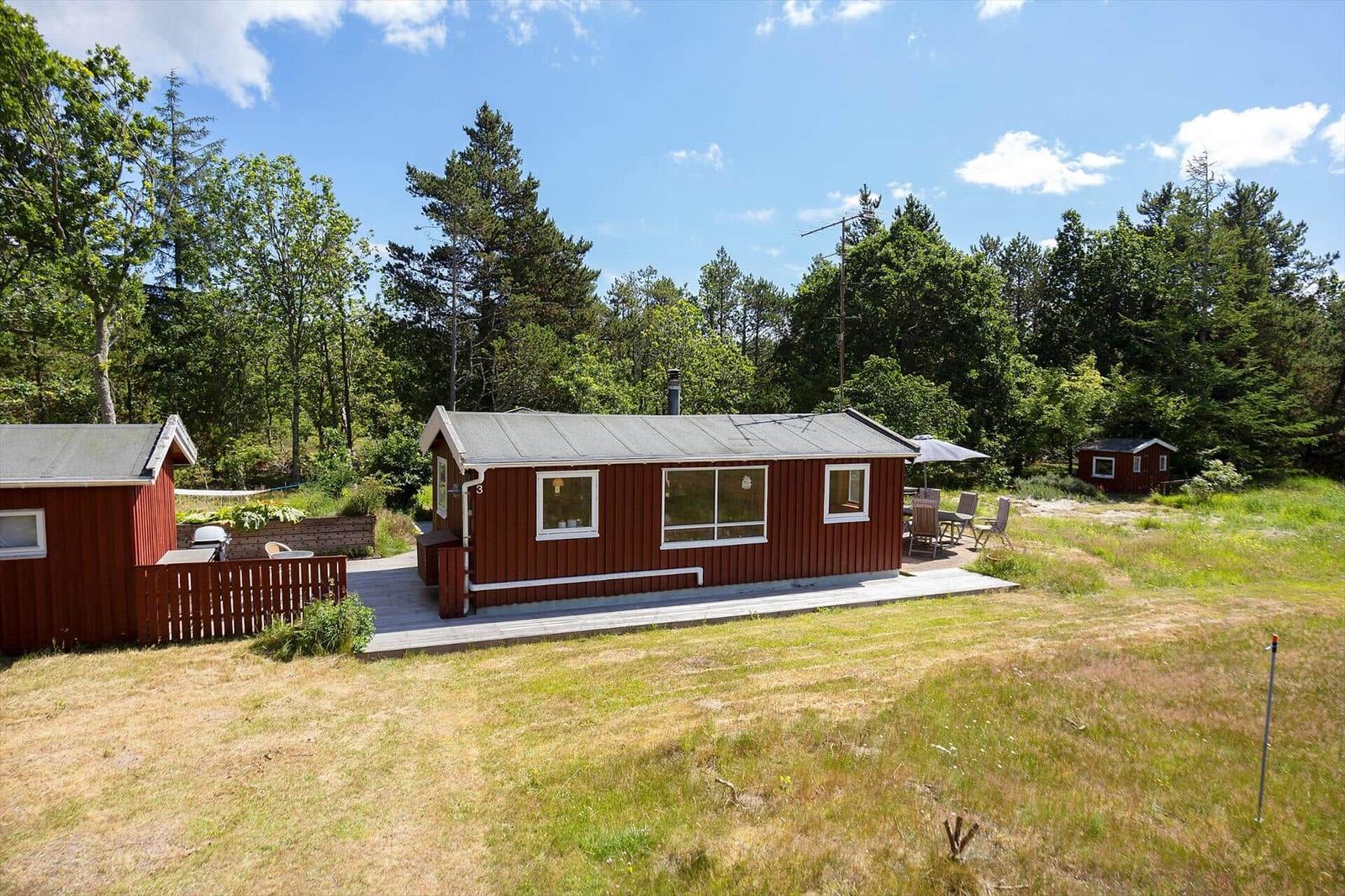 Rotes Holzhaus mit Terrasse und Garten. Umgeben von Bäumen und einem klaren Himmel.