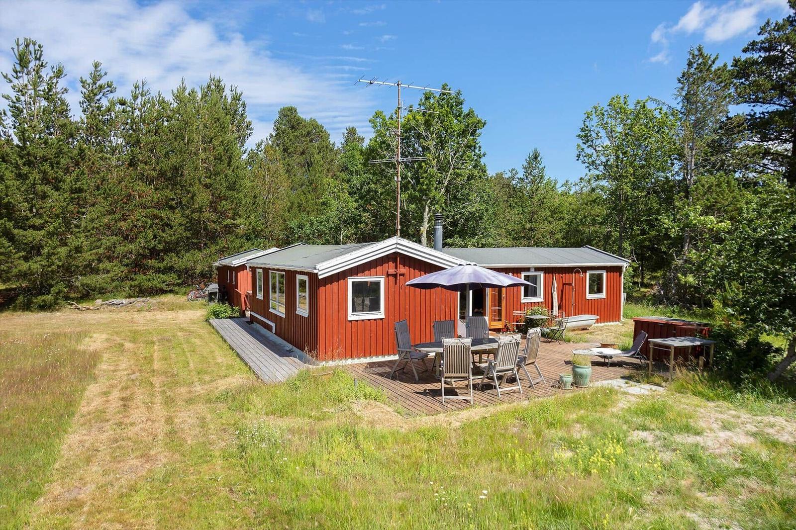 Roter Holzhaus mit Terrasse, Grill und Garten. Umgeben von Bäumen unter blauem Himmel.