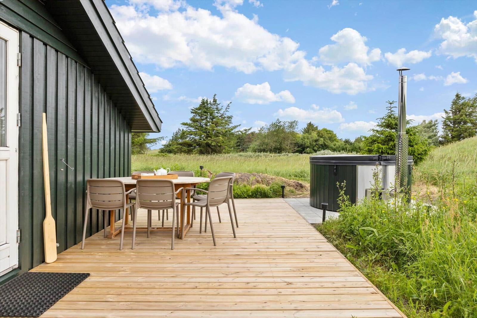 Holzterrasse mit Tisch und Stühlen neben einem Haus. Im Hintergrund liegt eine Wiese mit einem Whirlpool.