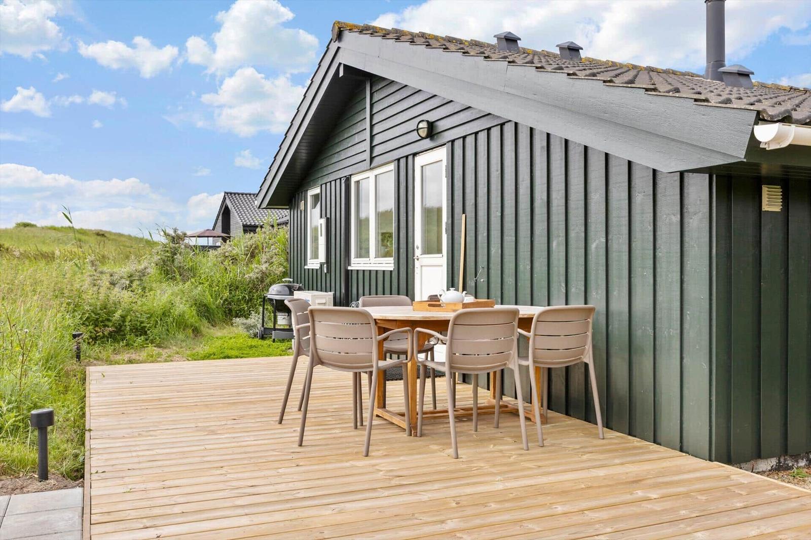 Holzterrasse mit Tisch und Stühlen vor einem grauen Holzhaus mit Dach und Fenstern.
