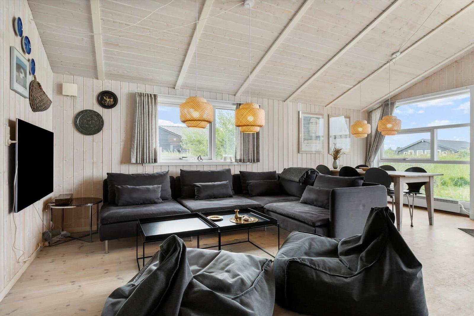 Wohnzimmer mit grauem Sofa, Tisch und Sitzgelegenheiten. Holzwände und Decke. Fenster mit Blick nach draußen.