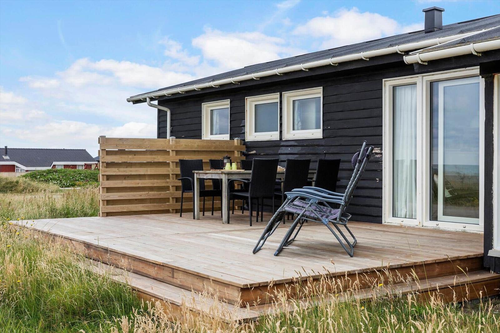 Holzterrasse mit Tisch, Stühlen und Liegestuhl vor einem schwarz getünchten Haus.