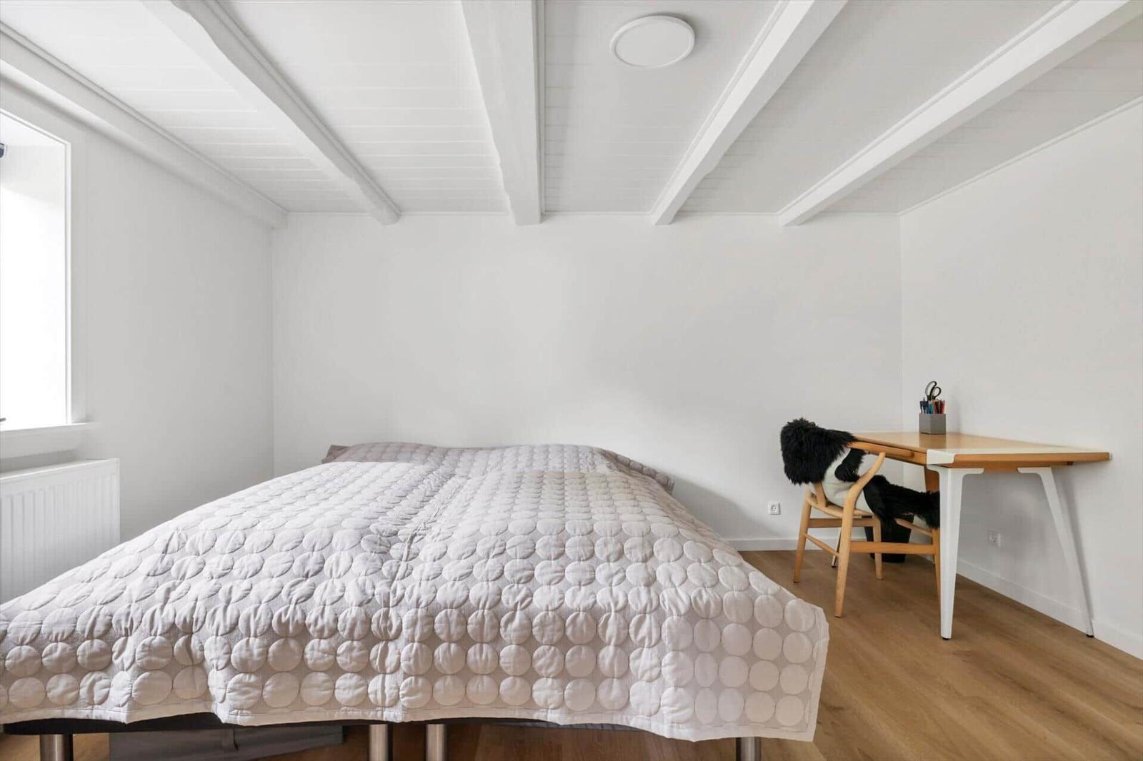 Schlafzimmer mit Bett, Schreibtisch und Fenster. Holzfußboden und weiße Wände.