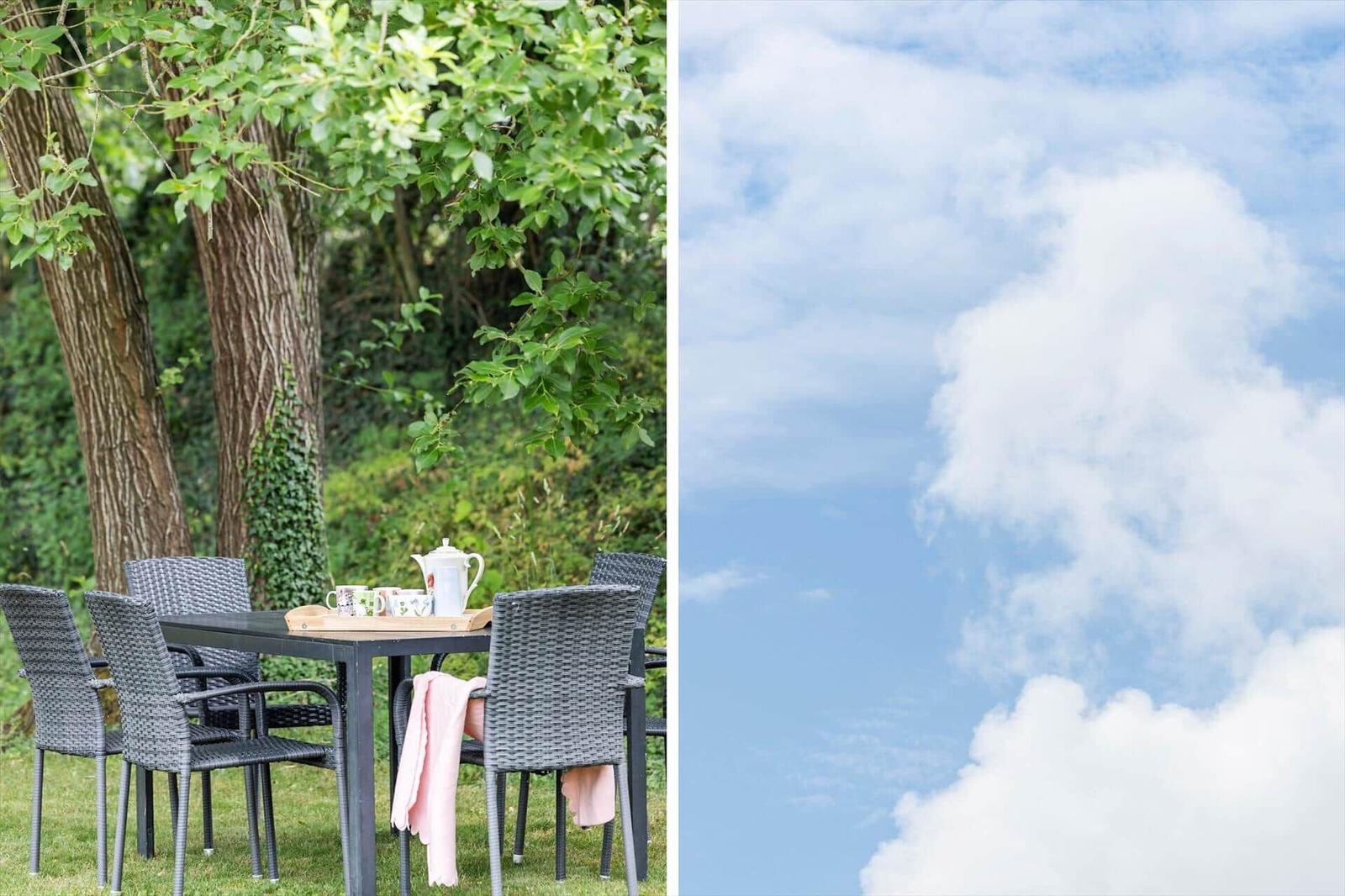 Außentisch mit Stühlen unter Bäumen und blauem Himmel mit weißen Wolken.