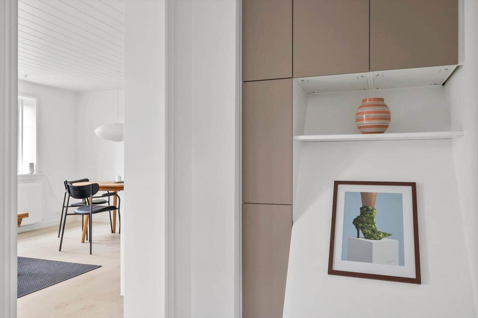 Ein Raum mit Holztisch, Stühlen und einer weißen Decke. An der Wand hängt ein Bild und ein Vase auf einer Ablage.
