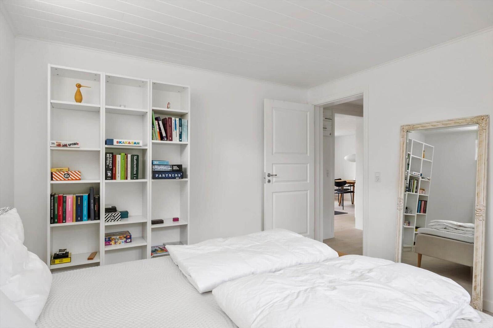 Schlafzimmer mit Bett, Bücherregal und Spiegel. Tür führt in einen weiteren Raum.