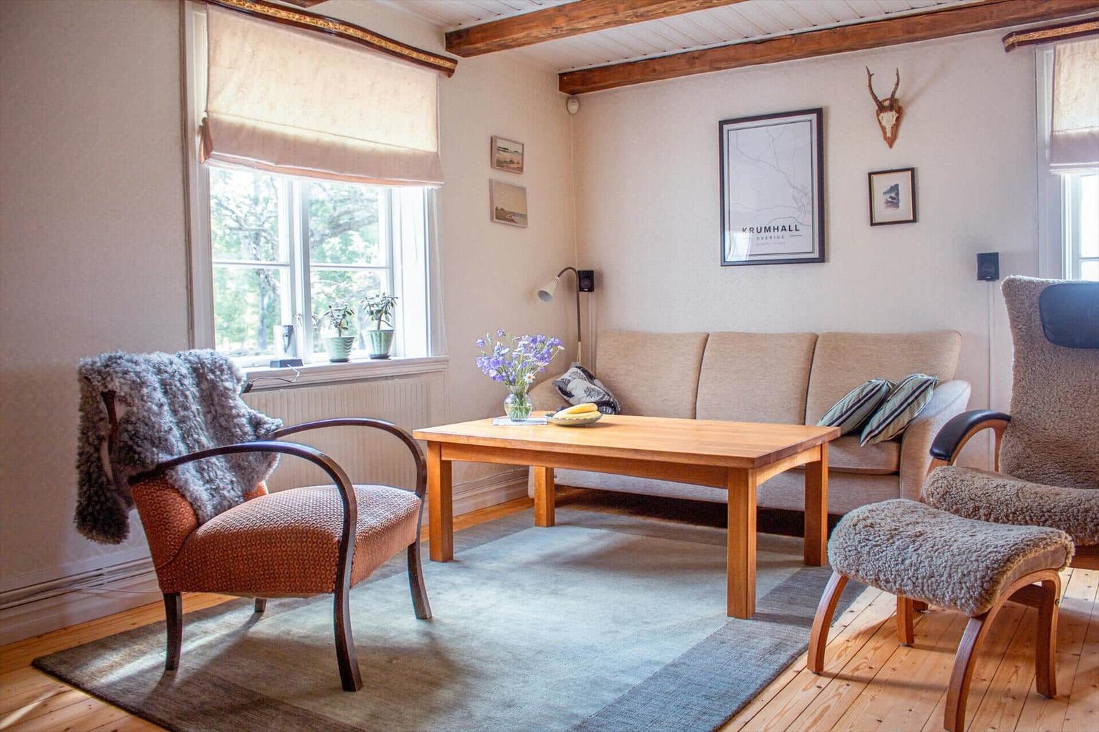 Wohnzimmer mit Sofa, Tisch und Stühlen. Holzbalken an der Decke und Fenster mit Blick nach draußen.