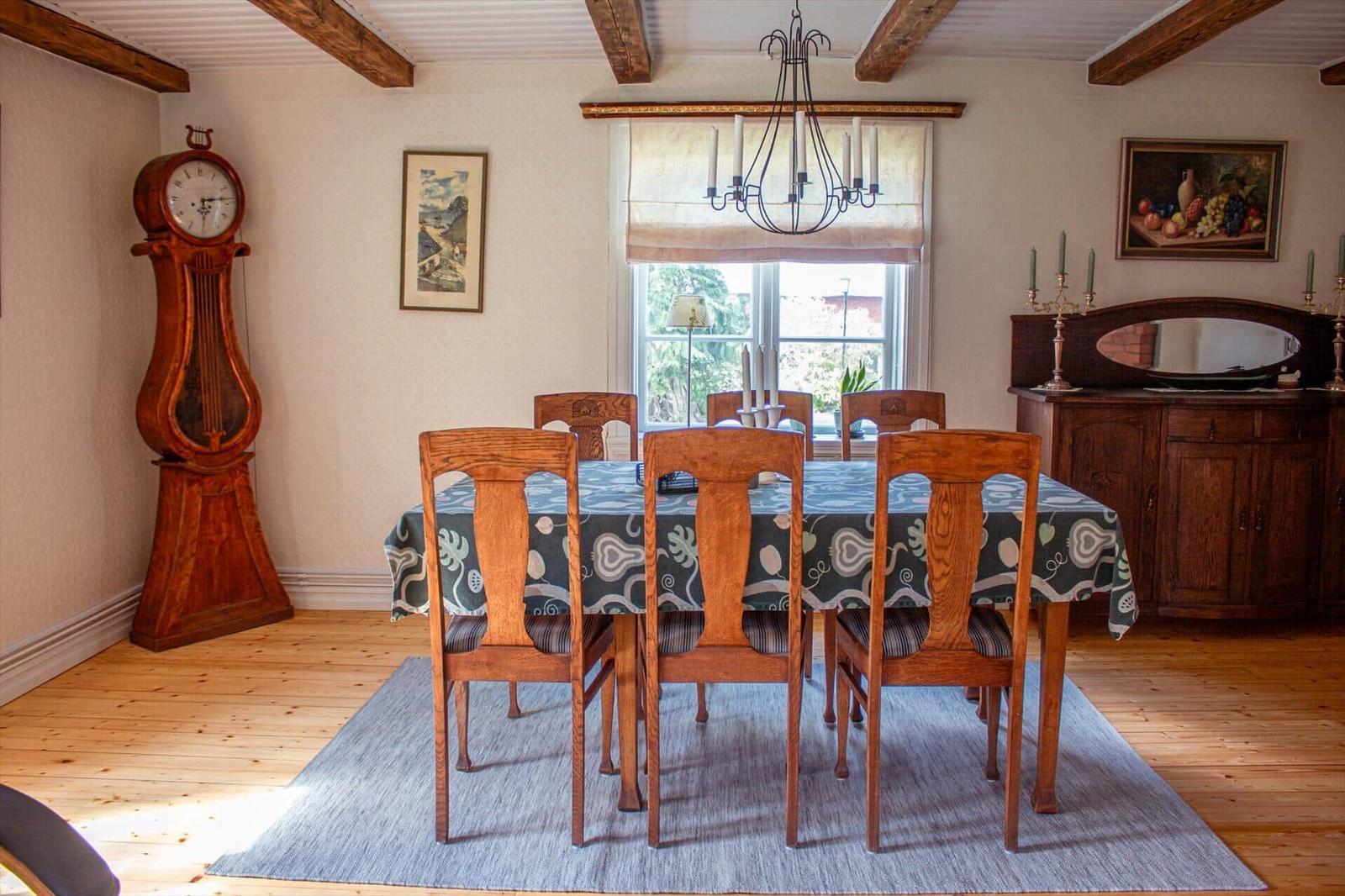 Esszimmer mit Holztisch, Stühlen, Tischdecke und Kronleuchter. An der Wand hängen Bilder und ein großer Wecker.