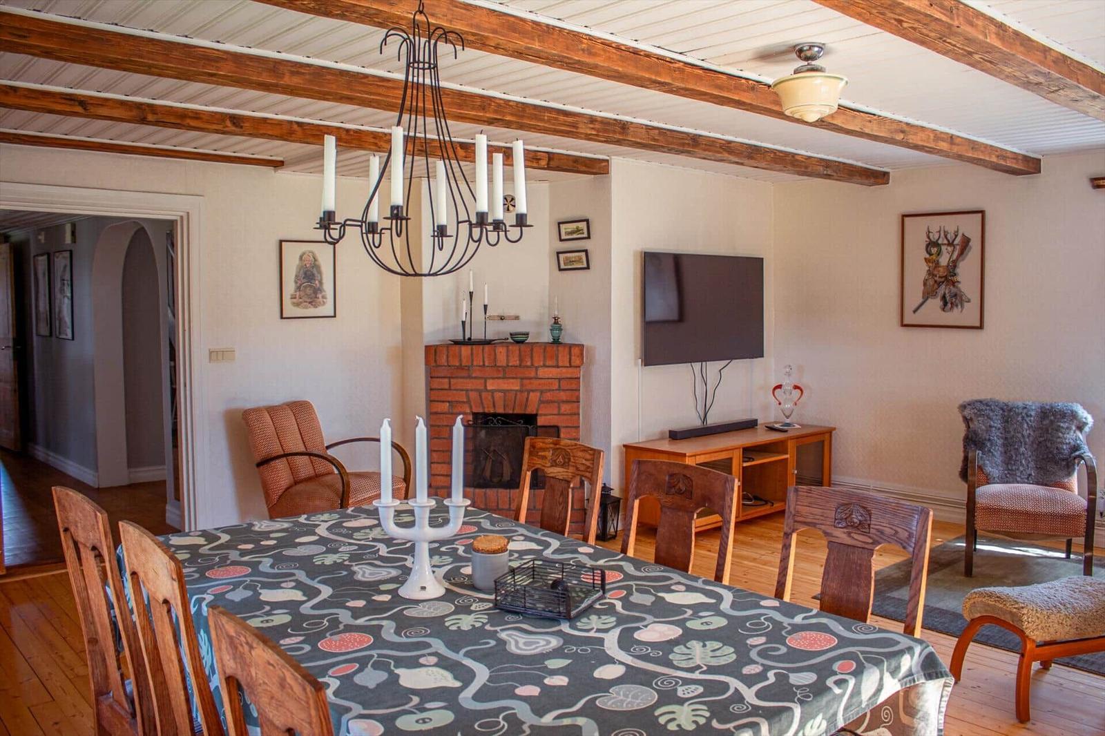 Esszimmer mit Holztisch, Stühlen und Kamin. Deckenbalken und Fernseher sichtbar.