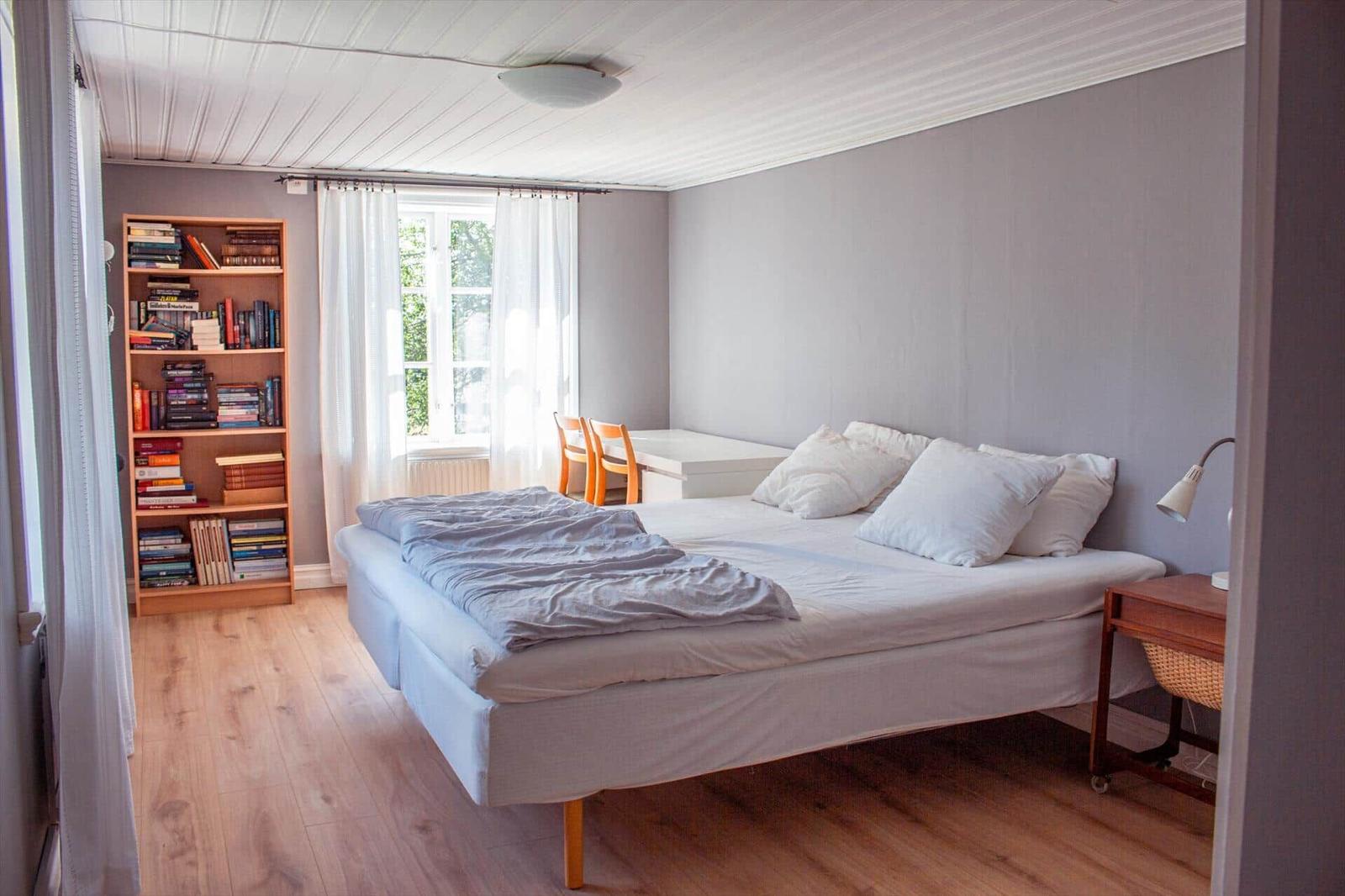 Schlafzimmer mit Bett, Bücherregal und Fenster. Holzboden und weiße Wände.