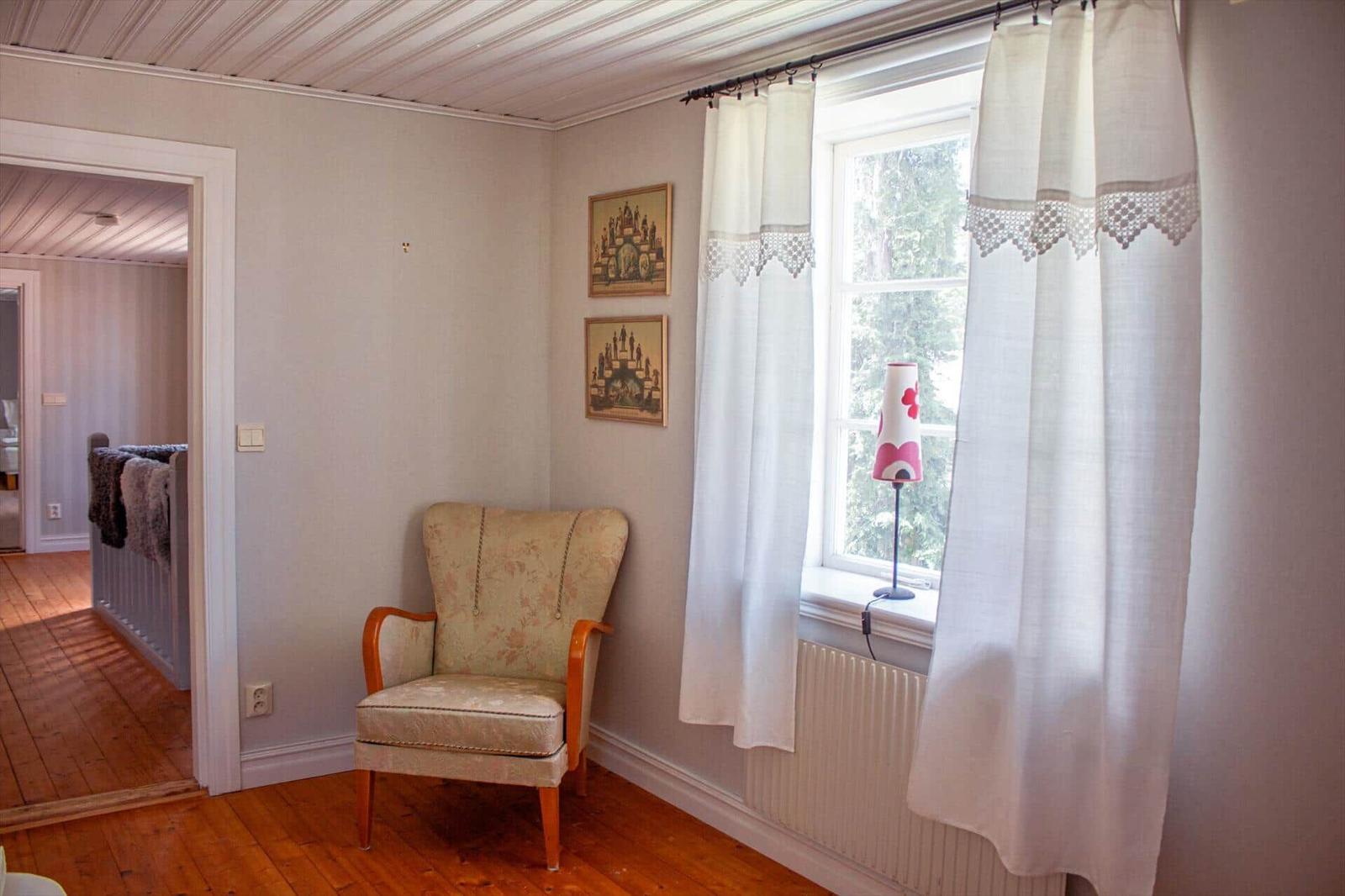 Ein Raum mit einem Sessel, einem Fenster mit weißen Vorhängen und zwei Bildern an der Wand.