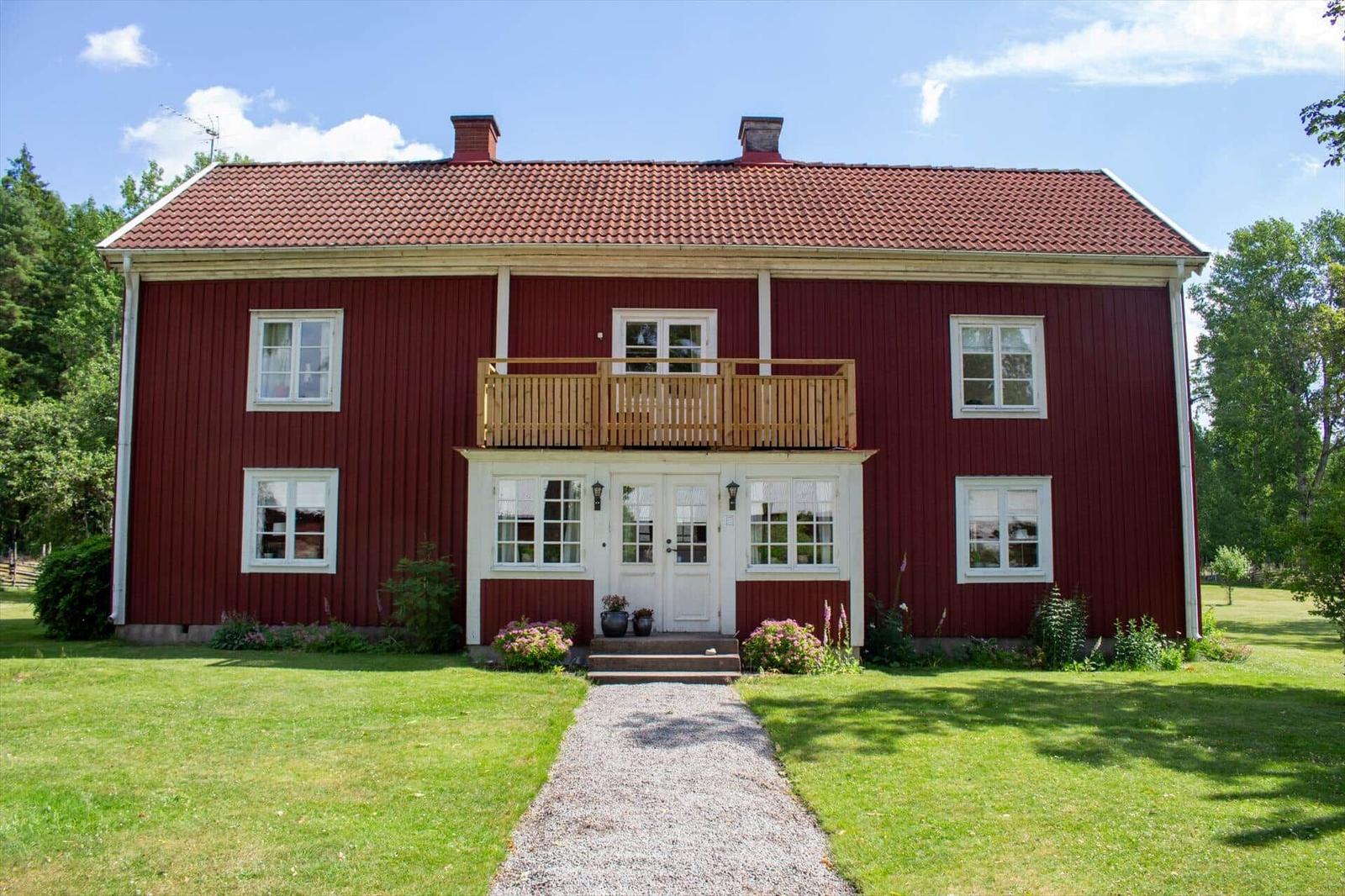 Rotes Holzhaus mit Balkon, weißen Fenstern und Eingang, umgeben von Gras und Bäumen.