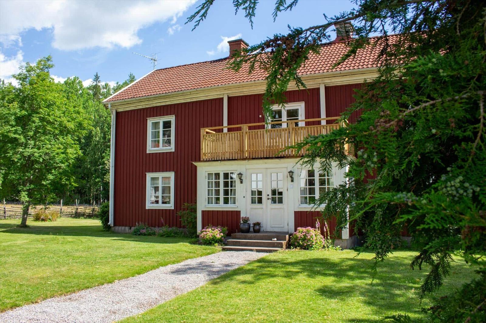 Rotes Haus mit Balkon, weißen Fenstern und Eingangstür. Grünflächen und Bäume umgeben das Gebäude.