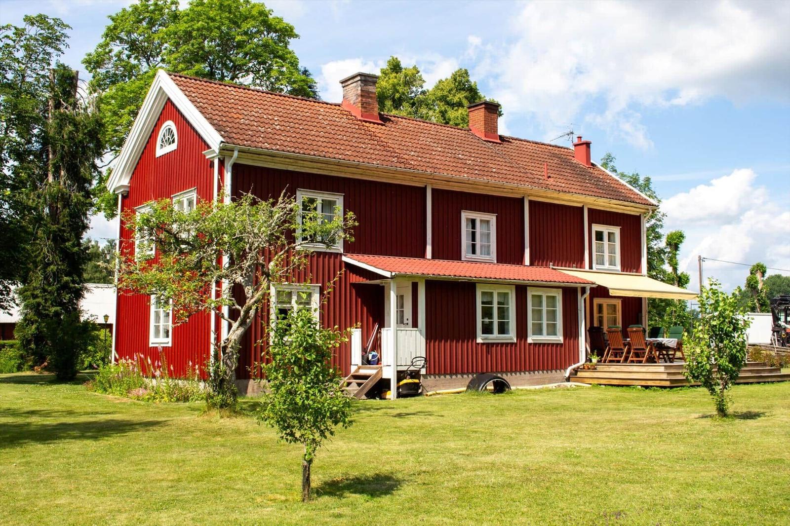 Rotes Holzhaus mit Terrasse und Garten. Mehrere Fenster und Schornsteine sind sichtbar.